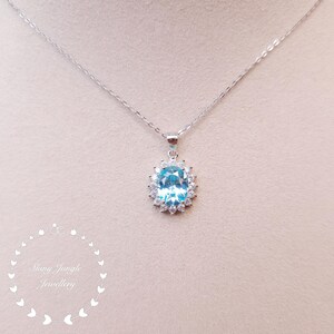 May include: A silver necklace with a pendant featuring a large, oval, light blue gemstone surrounded by smaller, clear gemstones. The pendant hangs from a delicate silver chain. The text "Shiny Jungle Jewellery" is visible.