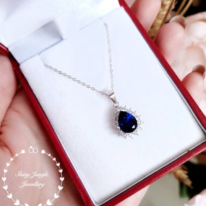 Puede incluir: Un collar de plata con un colgante de piedra preciosa azul en forma de pera rodeado de piedras preciosas transparentes m&aacute;s peque&ntilde;as. El collar est&aacute; en una caja de joyer&iacute;a roja.