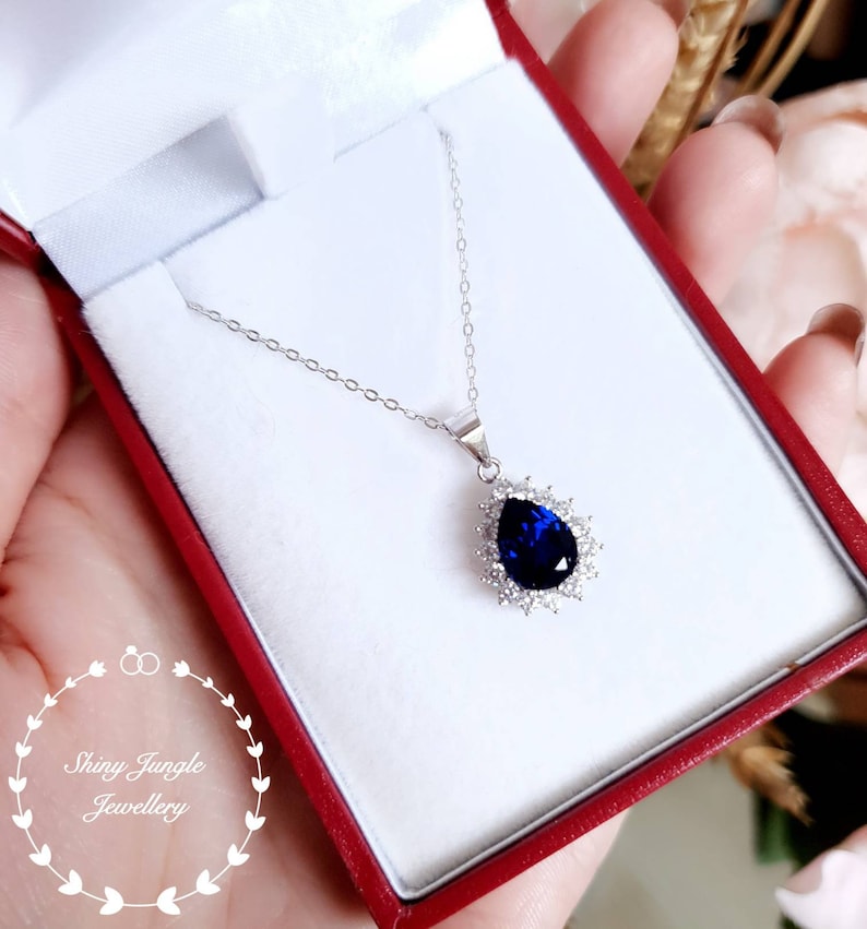 Puede incluir: Un collar de plata con un colgante de piedra preciosa azul en forma de pera rodeado de piedras preciosas transparentes m&aacute;s peque&ntilde;as. El collar est&aacute; en una caja de joyer&iacute;a roja.