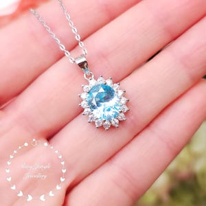 May include: A silver necklace with a pendant featuring a large, oval-shaped, light blue gemstone surrounded by smaller, clear gemstones. The pendant is suspended from a delicate silver chain. The image also includes the text "Shiny Jungle Jewellery".