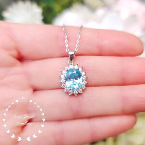 May include: A silver necklace with a large, oval, light blue gemstone pendant surrounded by smaller, clear stones. The pendant hangs from a delicate chain. The background is blurred, with the text "Shiny Jungle Jewellery" in a circular design.