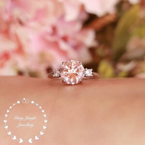 May include: A delicate engagement ring with a large, round, light pink gemstone surrounded by three smaller, round, clear gemstones. The ring is set in silver.