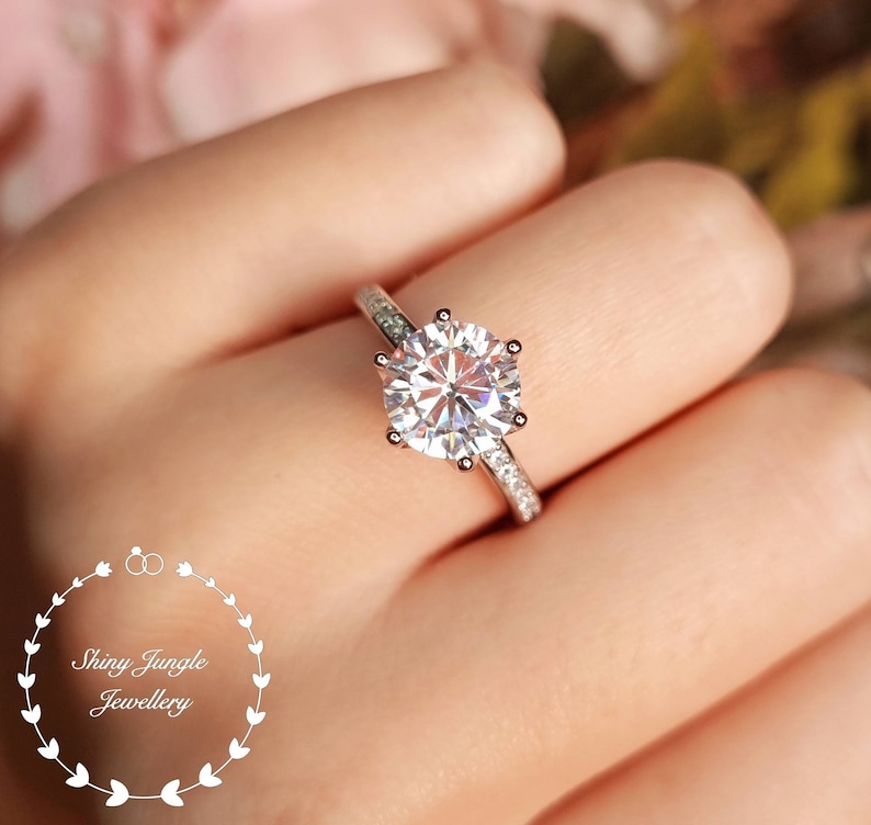 May include: A silver-toned ring featuring a large, round, brilliant-cut clear center stone held by six prongs, with smaller clear stones embedded along the band. The text Shiny Jungle Jewellery is visible in the lower left corner.