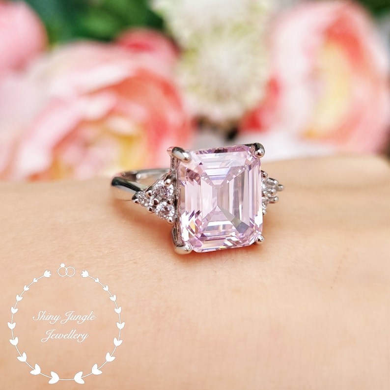 Pu&ograve; includere: Un anello d'argento con una grande pietra preziosa rosa quadrata e pietre preziose pi&ugrave; piccole e chiare sulla fascia.