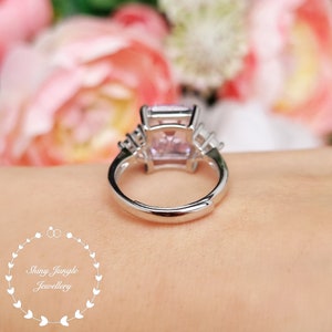 Pu&ograve; includere: Un anello d'argento con una grande pietra preziosa rosa di forma quadrata circondata da piccole pietre preziose chiare. L'anello &egrave; su un dito.