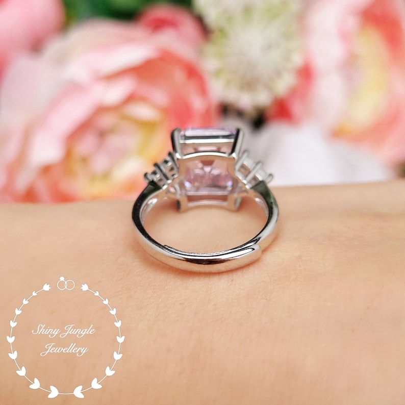 Pu&ograve; includere: Un anello d'argento con una grande pietra preziosa rosa di forma quadrata circondata da piccole pietre preziose chiare. L'anello &egrave; su un dito.