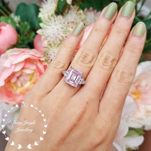 Pu&ograve; includere: Un anello d'argento con una grande pietra preziosa rosa a taglio smeraldo circondata da piccole pietre preziose chiare. L'anello &egrave; su un dito.