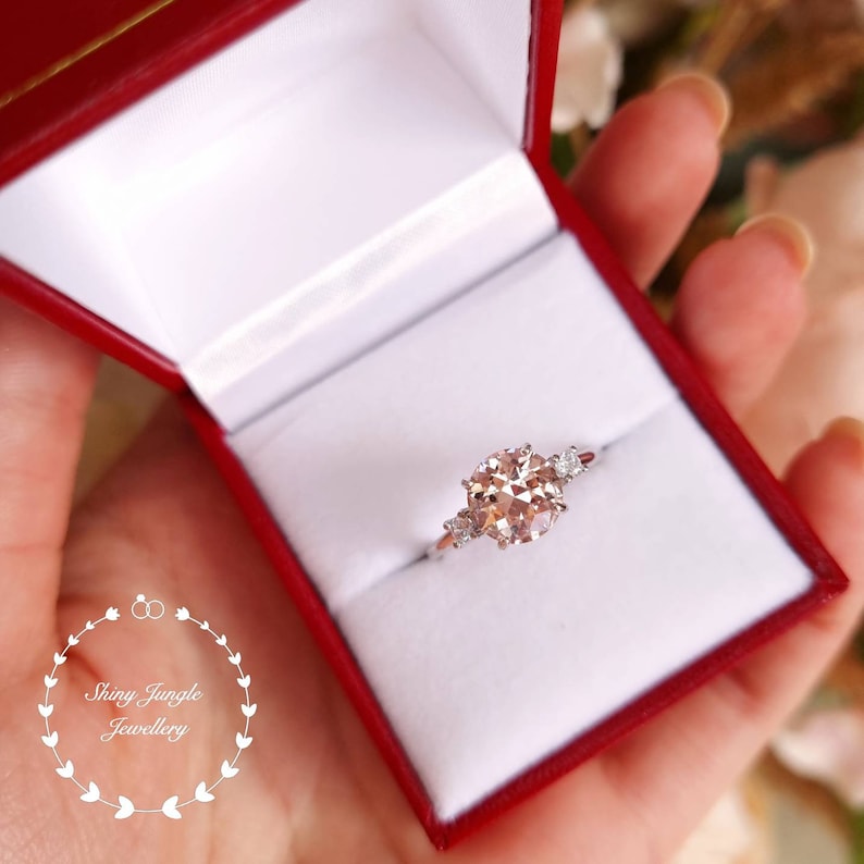 May include: A silver ring with a large, round, light pink gemstone surrounded by three smaller, round, white gemstones. The ring is in a red jewelry box.