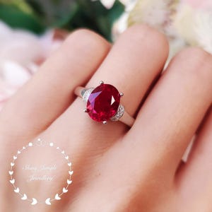 Könnte beinhalten: Ein silberfarbener Ring mit einem großen, oval geschliffenen, tiefroten Edelstein, der von vier Krappen gehalten wird. Die Ringschiene ist auf beiden Seiten der Fassung mit kleinen, klaren, funkelnden Steinen besetzt. Text Shiny Jungle Jewellery.