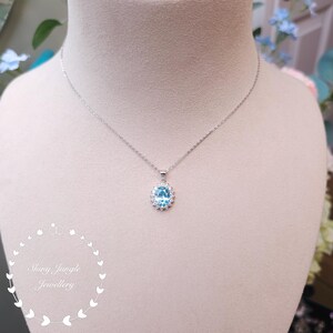 May include: A silver necklace with a pendant featuring a light blue oval gemstone surrounded by a halo of smaller clear stones. The necklace is displayed on a mannequin. The text "Shiny Jungle Jewellery" is in the lower left corner.
