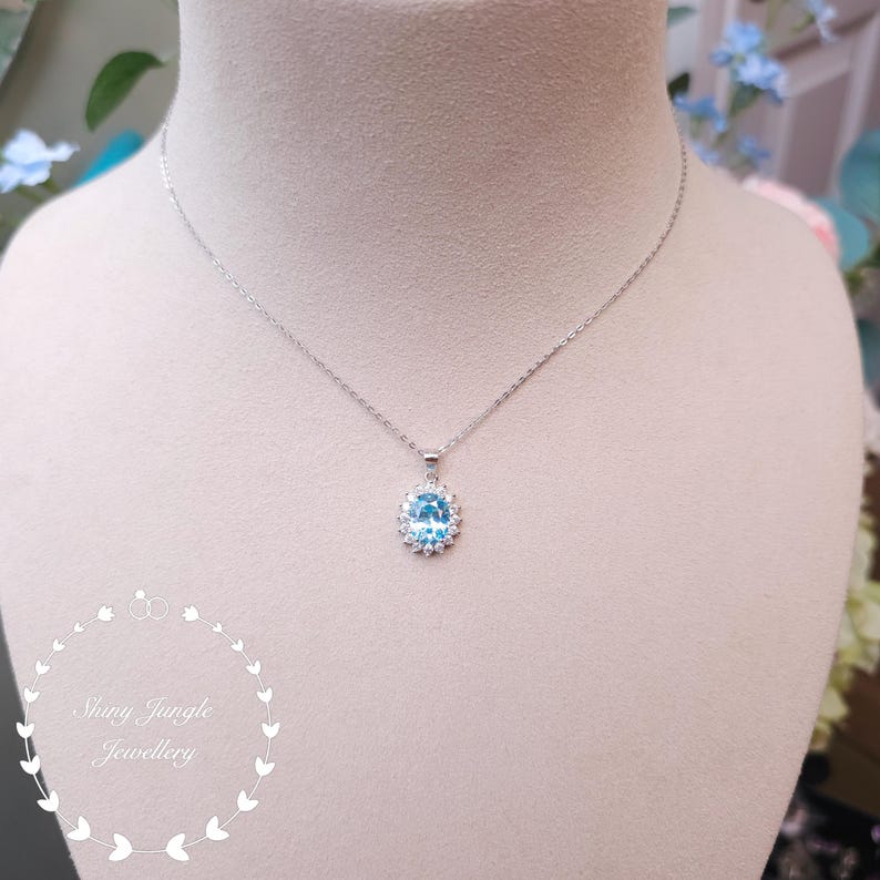 May include: A silver necklace with a pendant featuring a light blue oval gemstone surrounded by a halo of smaller clear stones. The necklace is displayed on a mannequin. The text "Shiny Jungle Jewellery" is in the lower left corner.