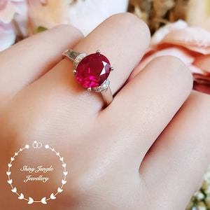 May include: A silver ring with a large oval-shaped ruby gemstone surrounded by smaller diamonds. The ring is on a person's finger.
