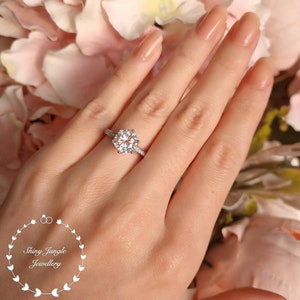 May include: A silver-toned ring featuring a large, round, brilliant-cut center stone held by six prongs, with smaller stones set along the band. The logo for Shiny Jungle Jewellery is visible in the lower left corner of the image.