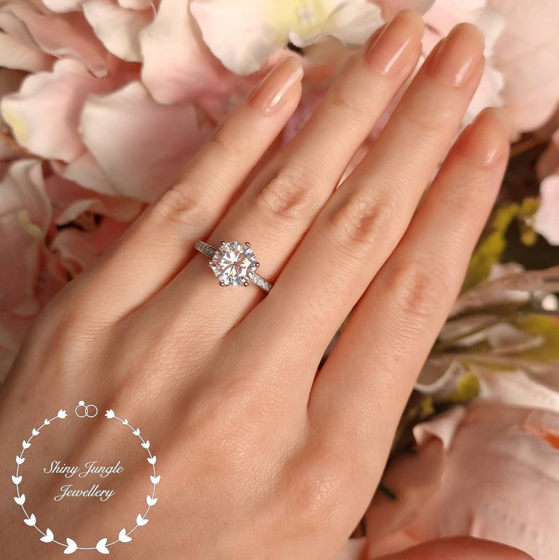 May include: A silver-toned ring featuring a large, round, brilliant-cut center stone held by six prongs, with smaller stones set along the band. The logo for Shiny Jungle Jewellery is visible in the lower left corner of the image.