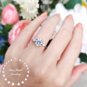 May include: A silver ring with a large round clear stone and two smaller clear stones on either side. The ring is on a person's finger.