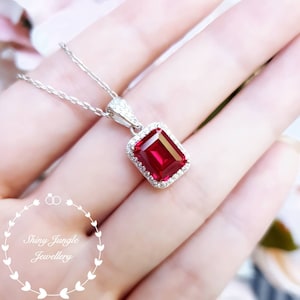 Puede incluir: Un collar de plata con un colgante de piedra preciosa roja de corte esmeralda rodeado de piedras preciosas transparentes más pequeñas. El colgante está engastado en un marco de plata.