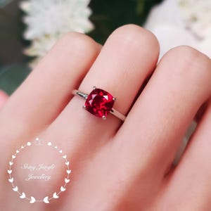 Puede incluir: Un anillo de plata con una gema roja brillante de talla cojín. El anillo tiene una banda delgada y pulida, y la gema está sujeta por cuatro garras. El anillo se muestra en un dedo, mostrando su elegante diseño. El logotipo de Shiny Jungle Jewellery está en la esquina inferior izquierda.