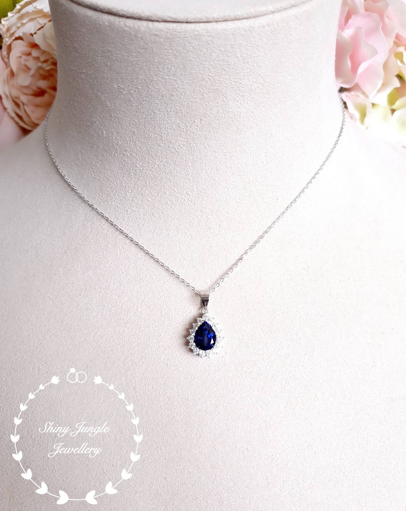 Puede incluir: Un collar de plata con un colgante en forma de l&aacute;grima que presenta una piedra preciosa azul oscuro rodeada de piedras preciosas transparentes m&aacute;s peque&ntilde;as.
