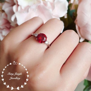 Könnte beinhalten: Ein silberner Ring mit einem großen roten Edelstein und zwei kleineren weißen Edelsteinen. Der Ring wird an einem Finger getragen.