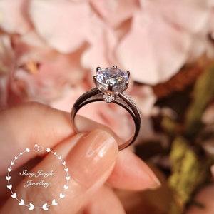 May include: A silver-toned ring featuring a large, round, faceted clear center stone held by a six-prong setting, with a smaller heart-shaped accent stone at the base of the setting and a band encrusted with tiny clear stones. Text reads: Shiny Jungle Jewellery.