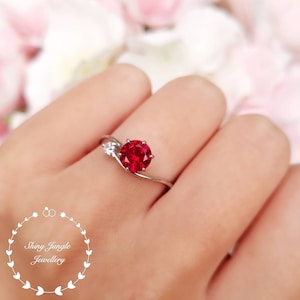 May include: A silver ring featuring a red gemstone and two smaller white gemstones. The ring is worn on a finger.