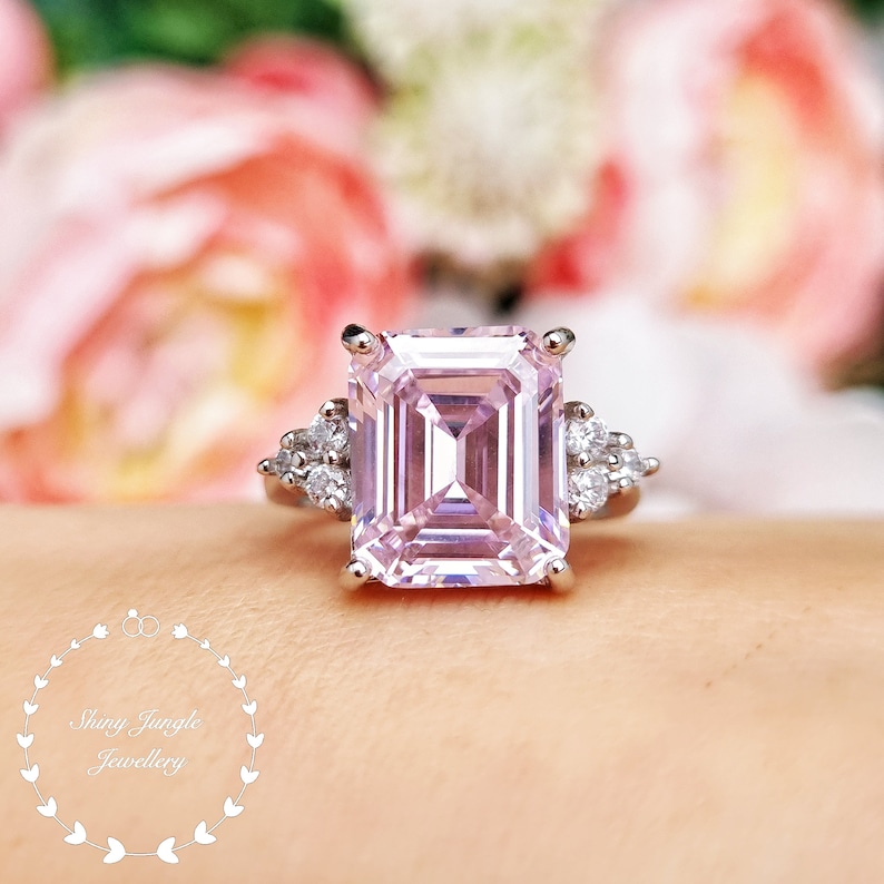Pu&ograve; includere: Un anello d'argento con una grande pietra preziosa quadrata di colore rosa chiaro circondata da pietre preziose pi&ugrave; piccole e trasparenti. L'anello &egrave; indossato su un dito.