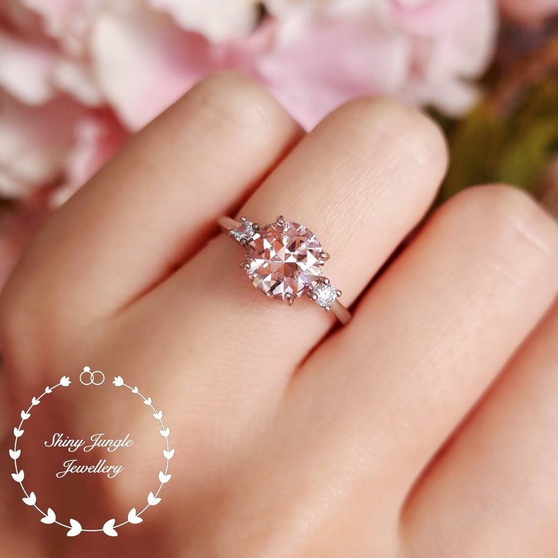 May include: A delicate engagement ring with a large, round, pale pink gemstone surrounded by smaller, clear gemstones. The ring is set in silver.