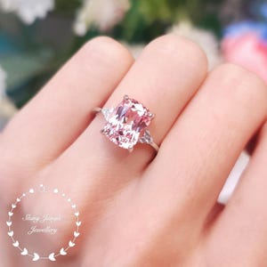 May include: A silver ring featuring a large, rectangular, light pink gemstone as the centerpiece. Two smaller triangular stones flank the main stone. The ring is on a finger, with the brand name "Shing Jungle Jewellery" in the lower left corner.