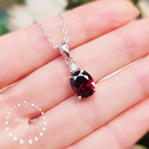 May include: A silver necklace with a pendant featuring a large, oval, deep red gemstone and a small, round, clear gemstone. The pendant hangs from a delicate silver chain. The necklace is displayed on a hand.