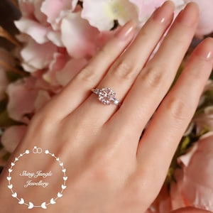 May include: A silver ring with a large, round, clear gemstone surrounded by smaller, clear gemstones. The ring is on a person's finger.