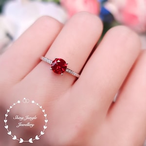 May include: A silver ring featuring a red gemstone surrounded by smaller clear gemstones. The ring is worn on a finger.