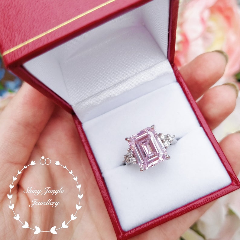 Pu&ograve; includere: Un anello di fidanzamento con taglio smeraldo rosa con piccoli accenti di diamanti incastonati in argento. L'anello &egrave; in una scatola per anelli in velluto rosso.