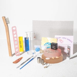 以下が含まれることがあります： 木製の歯ブラシ、ピンクの爪やすり、研磨布、小さな工具など、ジュエリーのクリーニングとケア用品のコレクション。画像にはイヤリング、ディスプレイスタンド、接着剤のチューブも含まれています。「Silver care basics」と書かれたカード。