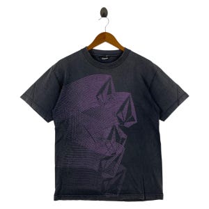 May include: Dark gray t-shirt with a purple geometric design. The graphic features a stylized, layered design. The shirt is hanging on a wooden hanger against a white background. The shirt has short sleeves and a crew neck.