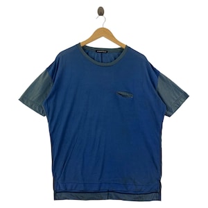 Può includere: Una t-shirt blu a maniche corte con maniche a contrasto in stile denim e una piccola tasca sul petto. La maglietta è appesa a una gruccia di legno su sfondo bianco. Lo scollo e l'orlo hanno un bordo blu scuro.