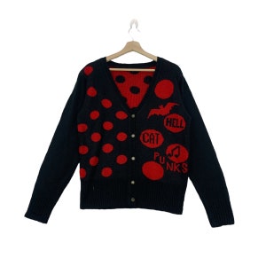 Op de afbeelding: Zwarte cardigan met rode stippen en rode accenten. De trui heeft een vleermuisafbeelding en de woorden "HELL", "CAT" en "PUNKS" in rode cirkels. Het is een ontwerp met knopen en een V-hals.