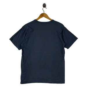 Pu&ograve; includere: Una maglietta blu navy appesa a una gruccia di legno. La maglietta a maniche corte &egrave; realizzata in un materiale morbido e ha uno scollo tondo. Il retro della maglietta &egrave; visibile, mostrando il colore pieno.