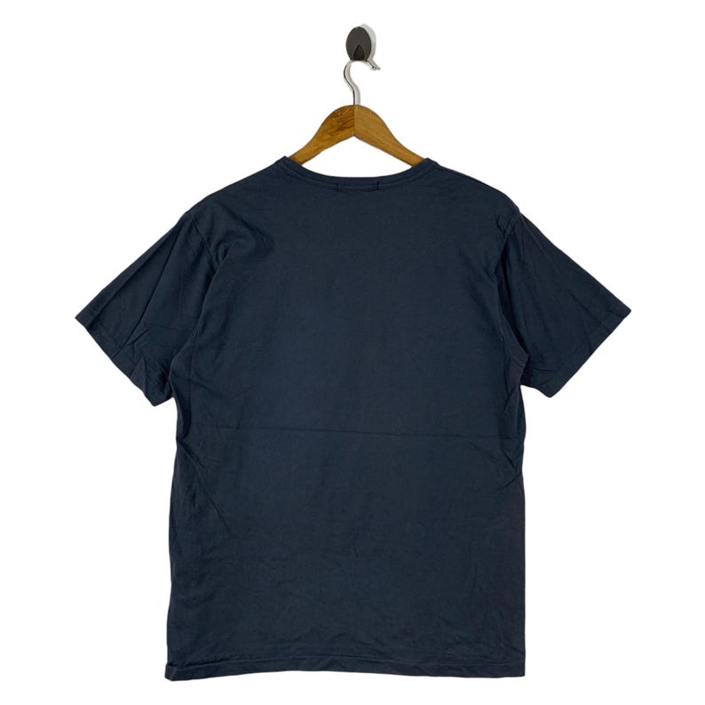 Pu&ograve; includere: Una maglietta blu navy appesa a una gruccia di legno. La maglietta a maniche corte &egrave; realizzata in un materiale morbido e ha uno scollo tondo. Il retro della maglietta &egrave; visibile, mostrando il colore pieno.