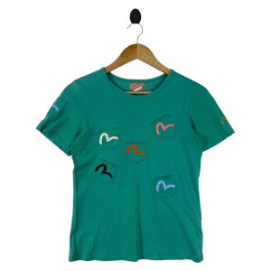 Puede incluir: Camiseta de color verde azulado con múltiples diseños de bolsillo con formas de "M" estilizadas y coloridas. La camiseta tiene cuello redondo y mangas cortas, y está colgada de una percha de madera.