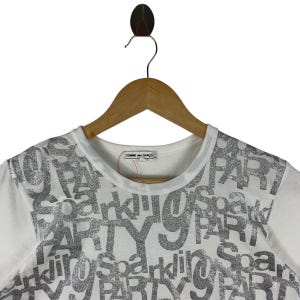 Puede incluir: Camiseta blanca con cuello redondo y mangas cortas. La camiseta presenta una impresi&oacute;n plateada brillante de las palabras "Sparkling Party" en un patr&oacute;n repetido. La etiqueta de la marca "Comme des Gar&ccedil;ons" es visible.