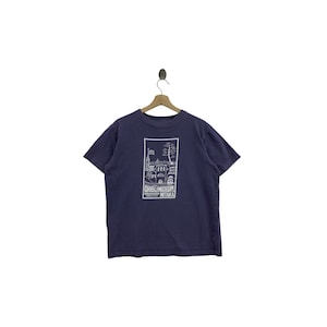 Puede incluir: Camiseta azul marino con un gráfico blanco de un edificio y árboles, y el texto "GHIBLI MUSEUM MITAKA" en un marco rectangular. La camiseta está colgada de una percha de madera.