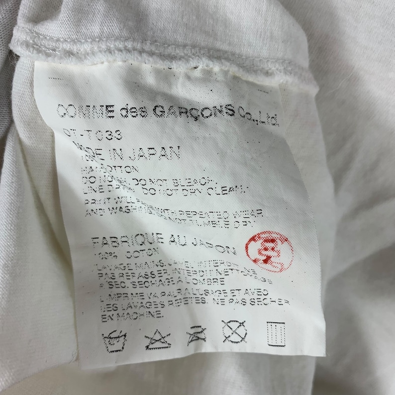 Puede incluir: Primer plano de una etiqueta de prenda blanca con texto negro. La etiqueta incluye la marca "COMME des GAR&Ccedil;ONS Co., Ltd." e instrucciones de cuidado. La etiqueta est&aacute; hecha en Jap&oacute;n y tiene un sello rojo.