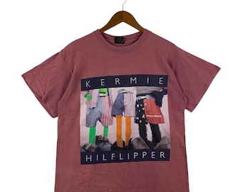 May include: A maroon t-shirt with a graphic print featuring the text "KERMIE" above a colorful image of figures wearing shorts and skirts. The text "HILFLIPPER" is printed below the image. The shirt is hanging on a wooden hanger.