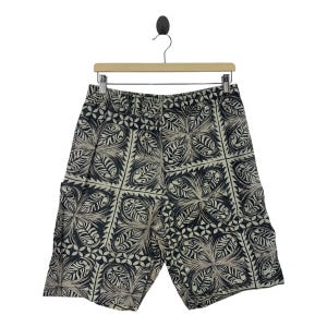 May include: Black and beige patterned shorts hanging on a wooden hanger. The shorts feature a repeating floral design. The shorts have an elasticated waistband and pockets. The shorts are made of a lightweight fabric.