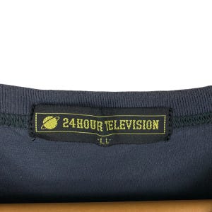 Pu&ograve; includere: Primo piano di una maglietta blu scuro con un'etichetta nera. L'etichetta presenta la scritta "24HOUR TELEVISION" in giallo, insieme a una grafica di un pianeta. &Egrave; visibile lo scollo della maglietta.