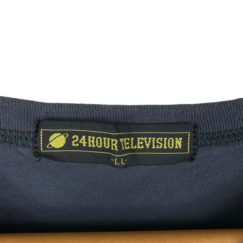 Pu&ograve; includere: Primo piano di una maglietta blu scuro con un'etichetta nera. L'etichetta presenta la scritta "24HOUR TELEVISION" in giallo, insieme a una grafica di un pianeta. &Egrave; visibile lo scollo della maglietta.