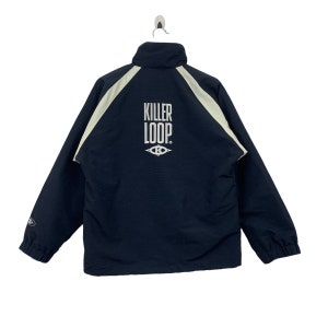 Op de afbeelding: Marineblauwe jas met een hoge kraag en witte accenten. De achterkant toont de witte tekst "KILLER LOOP" met een gestileerd oogsymbool. De jas hangt aan een metalen haak.