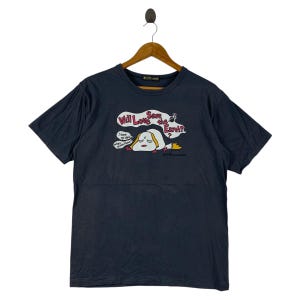 Pu&ograve; includere: T-shirt blu scuro con grafica a cartoni animati. La grafica presenta una creatura bianca e gialla con la scritta "Will Love Save the Earth?" e fumetti. La maglietta &egrave; appesa a una gruccia di legno.