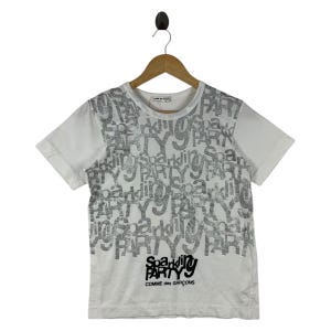 Puede incluir: Camiseta blanca con un dise&ntilde;o plateado brillante "Sparkling Party". La camiseta presenta el texto "Sparkling Party" y "Comme des Gar&ccedil;ons" en negro. La camiseta de manga corta est&aacute; colgada en una percha de madera.