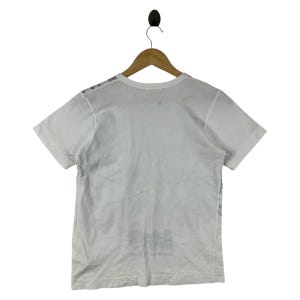 Puede incluir: Camiseta blanca de manga corta colgada de una percha de madera. La camiseta tiene cuello redondo y un dise&ntilde;o gr&aacute;fico en la parte inferior delantera. La camiseta est&aacute; hecha de un material suave y ligero.